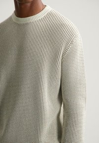 Prim-plan al unei persoane care poartă un pulover din tricot cu textură, de un bej deschis, cu un guler rotund și ribatat, pe un fundal simplu de culoare deschisă.