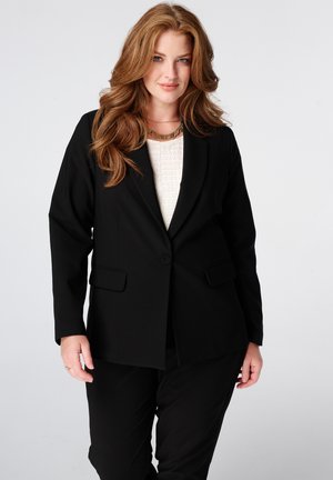 Vrouw met lang rood haar die een zwarte blazer en broek draagt over een wit topje, staand tegen een effen lichte achtergrond.