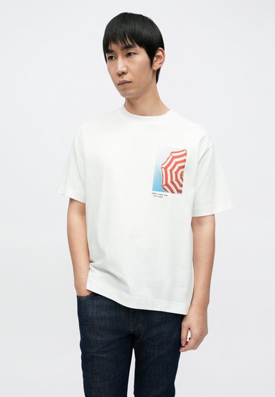 T-shirt en coton blanc avec un parapluie rayé rouge et blanc imprimé sur le côté gauche de la poitrine, accompagné d'un jean en denim bleu foncé.