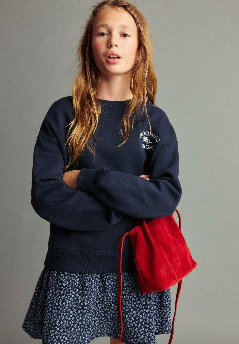 Mörkblå sweatshirt med vit text, blommönstrad kjol och en liten väska i röd mocka; långt vågigt hår med flätor.
