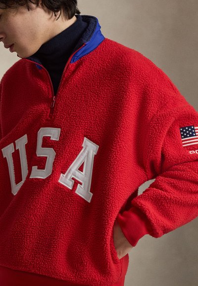 Νεαρό άτομο φορώντας κόκκινη φλις μπλούζα με γράμματα "USA" και πατρόν της αμερικανικής σημαίας στο μανίκι, με το χέρι στην τσέπη.