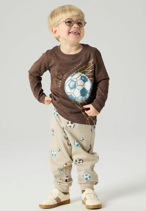 Bambino sorridente con occhiali, indossa una maglietta marrone a maniche lunghe con una grande grafica di un pallone da calcio, pantaloni beige con molteplici stampe di palloni da calcio e scarpe da ginnastica bianche.