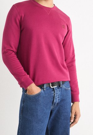 Mann trägt ein langärmliges dunkelrosa Sweatshirt mit Rundhalsausschnitt, blaue Jeans und schwarzen Gürtel, steht mit einer Hand in der Tasche.