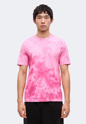 Mladý muž, ktorý nosí ružové tie-dye tričko s krátkym rukávom a čierne nohavice, stojaci pred jednoduchým svetlosivým pozadím.