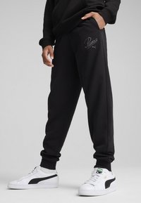 Czarne dresy wykonane z miękkiego materiału, z wyszywanym szarym logo Puma, elastycznymi mankietami i bocznymi kieszeniami, w zestawie z białymi sneakersami.