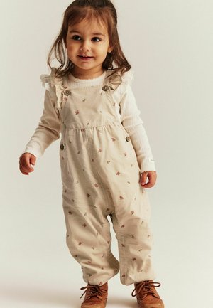 Hellbeige Cordoverall mit Blumenmustern, ausgestattet mit Rüschenträgern und Holzknöpfen, kombiniert mit einem weißen Langarmshirt und braunen Schuhen.