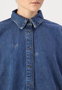 Camicia di denim blu con colletto, caratterizzata da bottoni argentati e fori a forma di cuore. Il tessuto presenta un aspetto testurizzato e cuciture visibili.