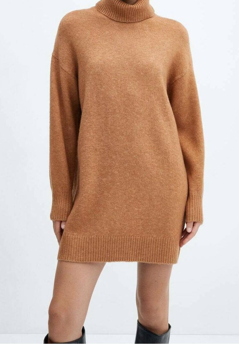 Femme portant une robe-pull oversize à col roulé couleur camel, associée à des bottes noires montantes, debout devant un fond blanc uni.