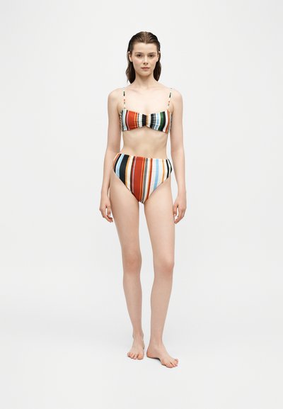 Jeune femme debout pieds nus portant un bikini à rayures verticales colorées avec un bas taille haute sur fond blanc.