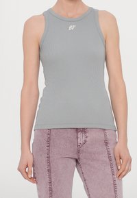 Jasnoszary prążkowany tank top z okrągłym dekoltem i małym białym logo na piersi, w połączeniu z jasnoróżowymi spodniami jeansowymi.