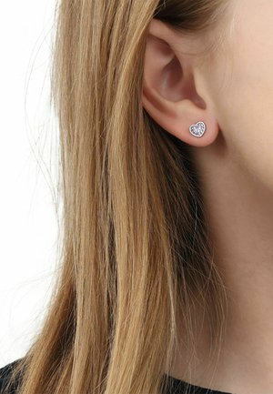Nahaufnahme eines Ohrs einer Person mit langen blonden Haaren, die einen kleinen silbernen herzförmigen Ohrstecker trägt.