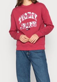 Osoba oblečená v červené mikině Tommy Jeans s bílým nápisem a modrých džínách, s rukama volně spojenýma na pase.