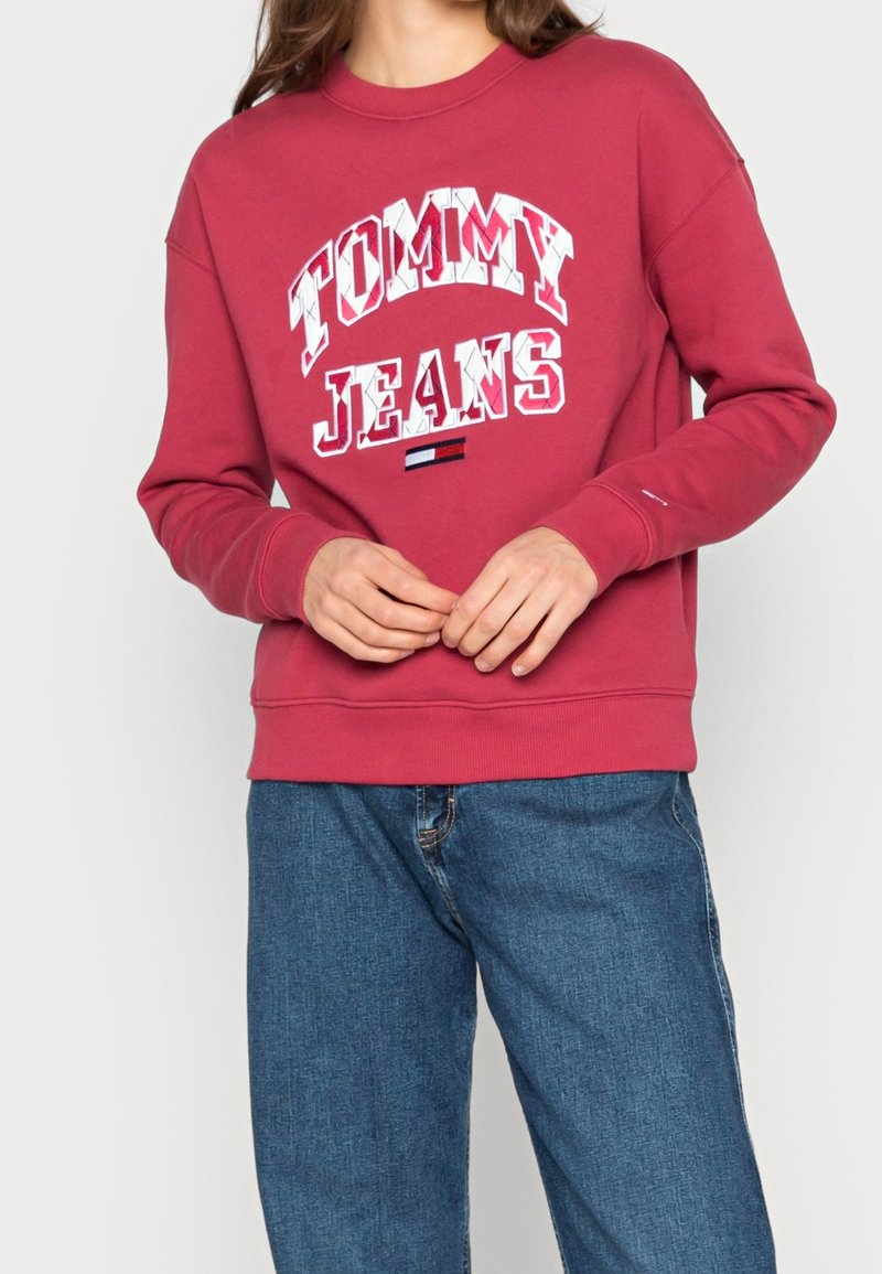 Osoba oblečená v červené mikině Tommy Jeans s bílým nápisem a modrých džínách, s rukama volně spojenýma na pase.