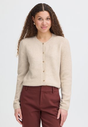Femme aux cheveux bouclés portant un cardigan beige boutonné et un pantalon bordeaux, debout devant un fond clair uni.