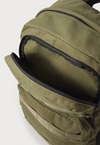 Mochila de color verde olivo hecha de un tejido duradero con cremalleras y una superficie texturada; cuenta con múltiples compartimentos y detalles de webbing táctico.