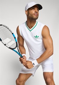Weißes ärmelloses Tennis-Shirt mit grünem Kragenbesatz, mit einem grünen Logo; weiße Shorts und Handgelenkbandagen. Hält einen Tennisschläger.