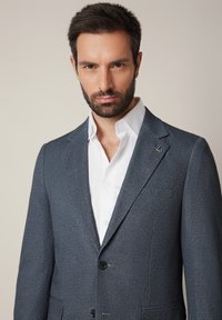 Szary żakiet teksturowany z klapami, dwoma przednimi kieszeniami i zapięciem na jeden guzik, noszony na białej koszuli.