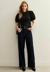 Maglione nero con texture e maniche a sbuffo, abbinato a pantaloni a gamba larga in denim scuro a vita alta e décolleté neri a punta.