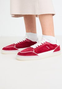 Sneaker rosse in suede con accenti bianchi, lacci e suola in gomma trasparente, caratterizzate da un design elegante e una brandizzazione sottile sul lato.