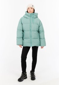 Chaqueta de plumas en color azul verdoso con cuello alto y cierre de cremallera, que presenta una forma cuadrada y un diseño acolchado. Llevada sobre pantalones negros y botas.