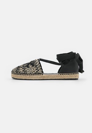 Espadrille noire et beige avec empeigne tissée, liens en ruban à la cheville et semelle en jute. Présente un bout arrondi et un talon noir lisse.