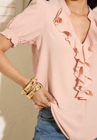 Světle růžová blůza s volány a krátkými nařasenými rukávy; doplněná zlatými náramky; jemná textura; design ve tvaru V. Viditelné casual džíny.