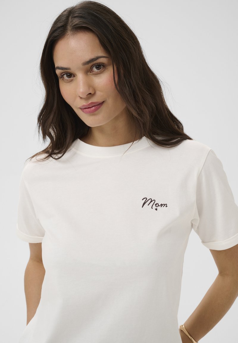 Femme aux longs cheveux foncés portant un T-shirt blanc avec « Maman » brodé sur la poitrine, posant devant un fond clair uni.