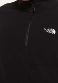 Pullover in pile nero con cerniera corta sul davanti e logo bianco "The North Face" sul lato sinistro del petto.