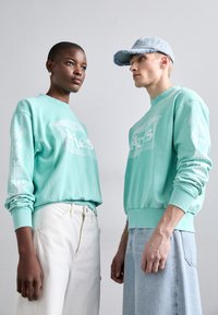 Bluze de molton verde mentă cu imprimeu "Berbec", cu mansete strâmte și croială lejeră, asortate cu pantaloni albi și bleu deschis. Pălăria are marginea sfâșiată.