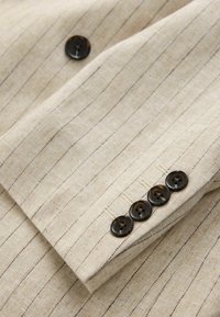 Manche de costume beige à fines rayures avec quatre boutons brun foncé cousus en ligne près du poignet.