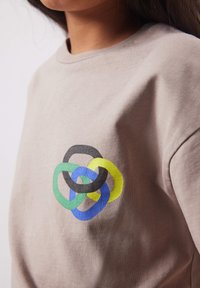 Personne portant une chemise beige avec un motif géométrique entrelacé coloré en vert, bleu, noir et jaune sur la poitrine.