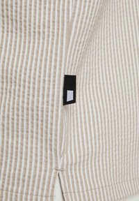 Tejido a rayas con líneas beige y blancas, que presenta una abertura lateral y una etiqueta negra con un acento cuadrado blanco. Textura suave.
