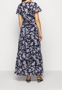 Kvinna i en lång marinblå klänning med vita och ljuslila blommönster, svävande ärmar och svarta klacksandaler, stående mot en vit bakgrund.