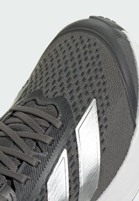 Zapatilla atlética gris con parte superior de malla texturizada, cordones oscuros, suela de goma y acentos metálicos plateados en el lateral.