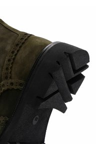 Bota de ante en verde oliva con un patrón de brogue decorativo y una suela de goma negra con un diseño texturizado y en ángulo para mayor agarre.