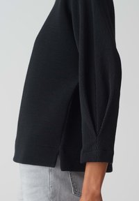 Haut noir à manches longues côtelé avec fentes latérales, offrant une coupe décontractée et un tissu texturé. Comprend des poignets roulés et un col haut.