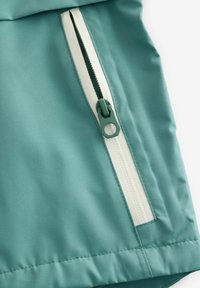 Gros plan d'une fermeture éclair blanche partiellement ouverte sur un tissu bleu sarcelle, montrant le curseur et les détails de la couture.