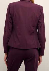 Paarse op maat gemaakte blazer met een gestructureerd ontwerp, lange mouwen en subtiele naden. Het materiaal lijkt glad en de stof heeft een lichte glans.