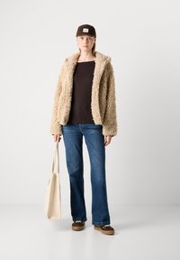 Kvinne i beige fleece-jakke, mørkebrun skjorte, blå jeans, brun caps og sneakers, som holder en beige totebag, står foran en hvit bakgrunn.
