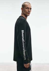 Długa czarna koszula z bawełny, z okrągłym dekoltem i białym nadrukiem na lewym rękawie. Gładka tekstura, luźny fason.