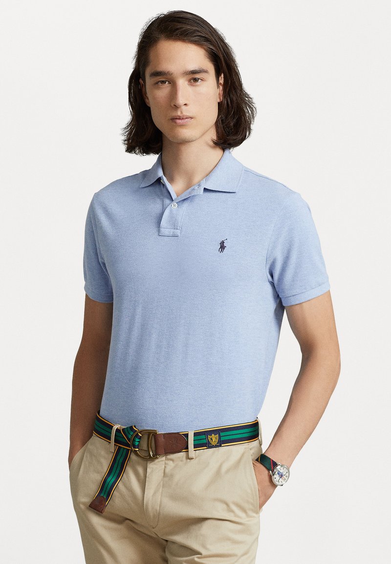 Polo de culoare albastru deschis, cu mânecă scurtă, dotat cu un guler cu două nasturi și un logo brodat. Combinat cu pantaloni kaki și o curea cu dungi multicolore.