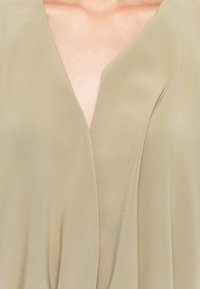 Gros plan sur une personne portant un vêtement beige à col en V en tissu doux, avec des plis subtils autour de la poitrine.