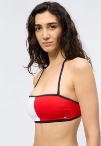 Γυναίκα με μακριά, σγουρά μαλλιά που φοράει κόκκινο και λευκό στρωτό μπικίνι τοπ τύπου bandeau με λεπτές ναυτικές μπλε τιράντες.