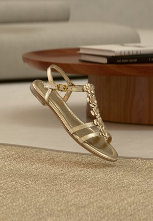 Χρυσό σανδάλι με τρεις λεπτές λουρίδες, μια ανθοδέσμη στο μπροστινό μέρος και ένα ρυθμιζόμενο λουρί στον αστράγαλο με μεταλλική αγκράφα.