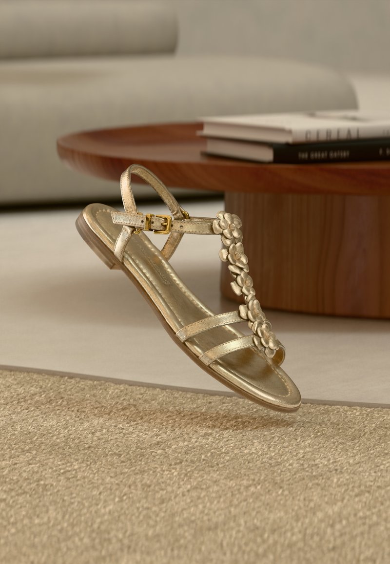 Χρυσό σανδάλι με τρεις λεπτές λουρίδες, μια ανθοδέσμη στο μπροστινό μέρος και ένα ρυθμιζόμενο λουρί στον αστράγαλο με μεταλλική αγκράφα.