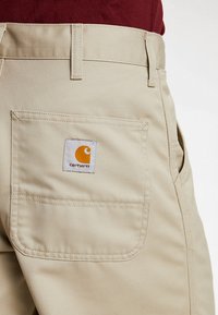 Beige Arbeitshose mit einer auffälligen Gesäßtasche, die ein quadratisches Carhartt-Logo in Orange und Weiß zeigt, aus strapazierfähigem Stoff mit glatter Textur.