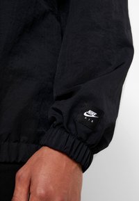 Černý nylonový rukáv bundy s elastickým manžetovým zakončením a obdélníkovou nášivkou Nike Air na zápěstí, s texturovaným povrchem.