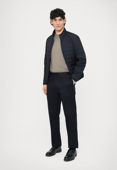 Temno modra prešita jakna preko svetlo sive teksturirane majice, kombinirana s temnimi hlačami in črnimi usnjenimi čevlji, stoji na belem ozadju.