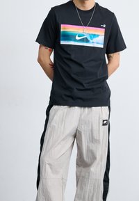 Černé tričko s krátkým rukávem s barevným motivem vlny a logem Nike uprostřed, doplněné světlými texturovanými kalhotami s pruhy na stranách.