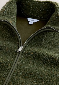Primo piano di giacca in pile verde oliva con cerniera argentata parzialmente aperta, che mostra la morbida texture e l'etichetta interna con la scritta "Next" e taglia M.